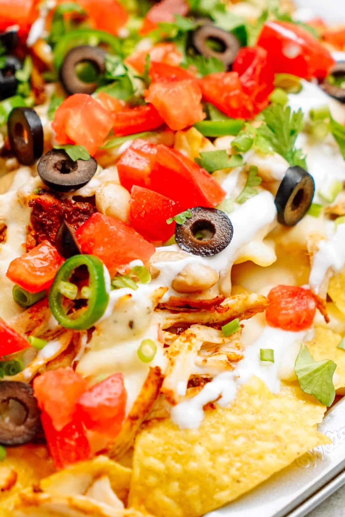 Close-up of nachos topped with diced tomatoes, black olives, green onions, jalapeños, shredded chicken, melted cheese, and a drizzle of white sauce.