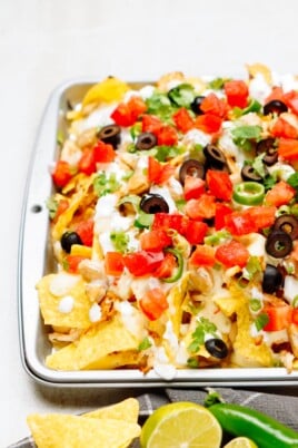 A tray of loaded nachos topped with cheese, tomatoes, black olives, green onions, and drizzled with sour cream, with lime wedges and a green chili pepper placed beside it.