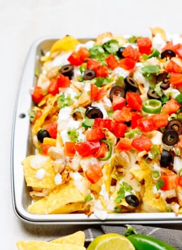 A tray of loaded nachos topped with cheese, tomatoes, black olives, green onions, and drizzled with sour cream, with lime wedges and a green chili pepper placed beside it.