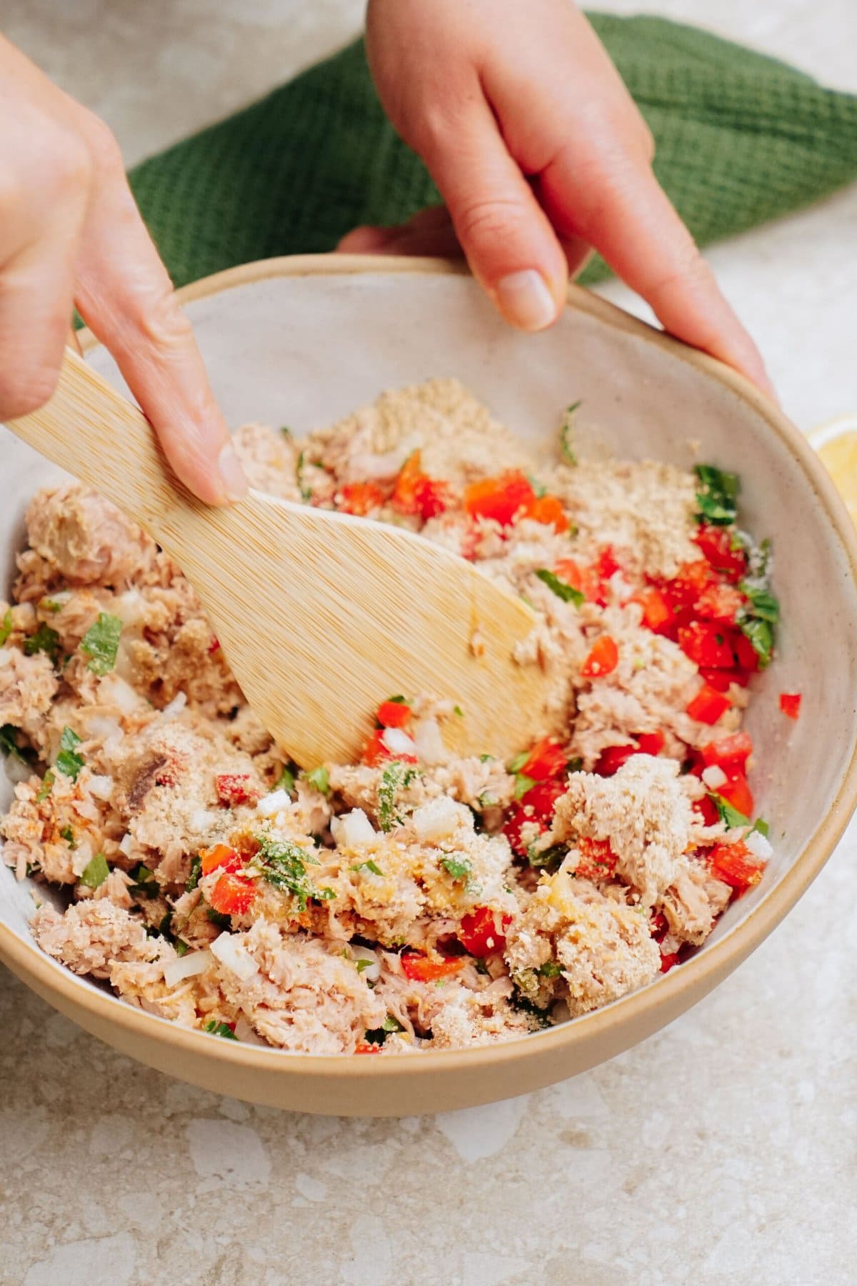 A person mixing a bowl of ground meat, chopped red bell peppers, herbs, and bread crumbs with a wooden spatula, preparing delicious tuna patties.