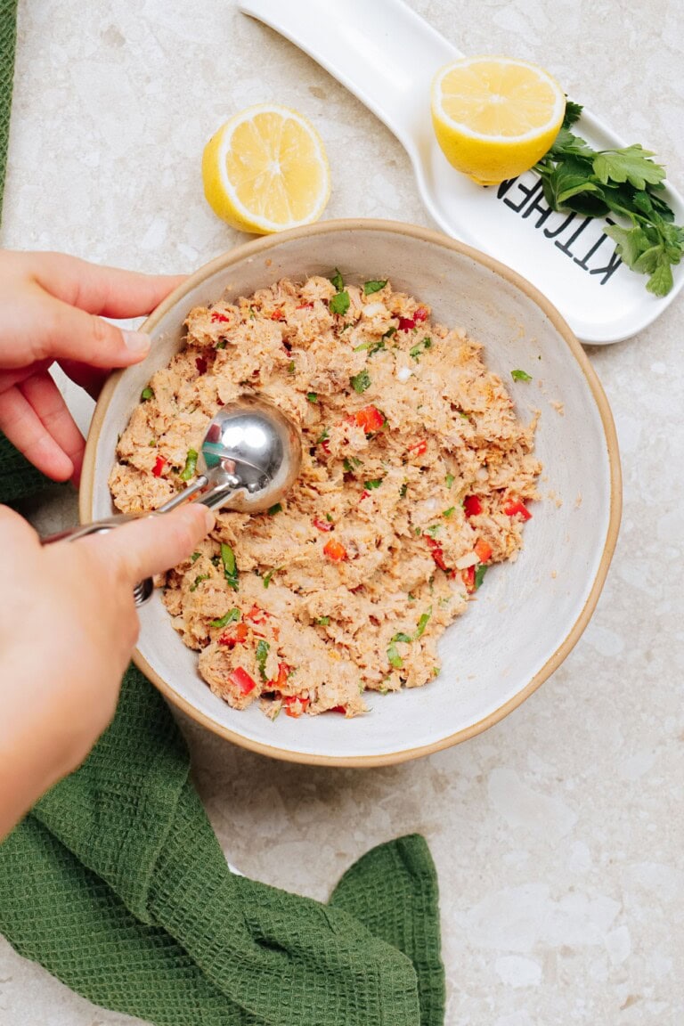 Hands using an ice cream scoop in a bowl of seasoned tuna mix on a counter with lemon halves, parsley, and a green cloth, preparing Tuna Patties.