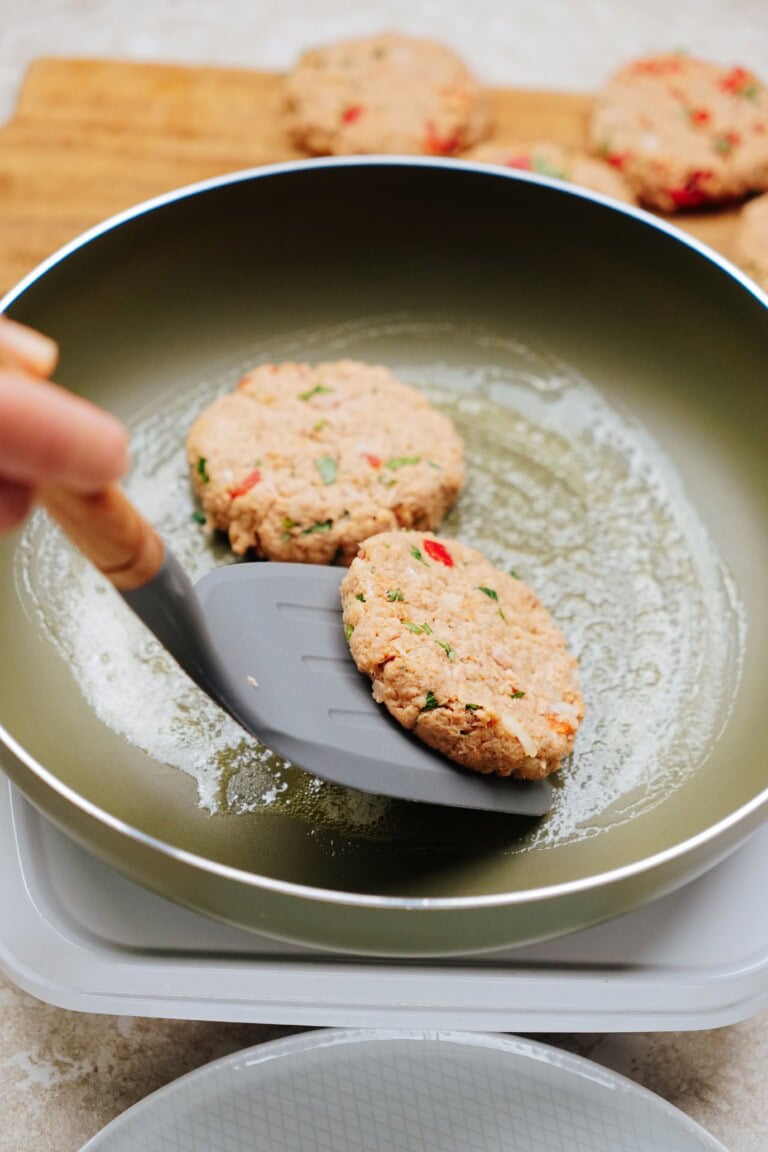 A hand is flipping a tuna patty with a spatula in a frying pan, while other uncooked tuna patties sit in the background on a cutting board.