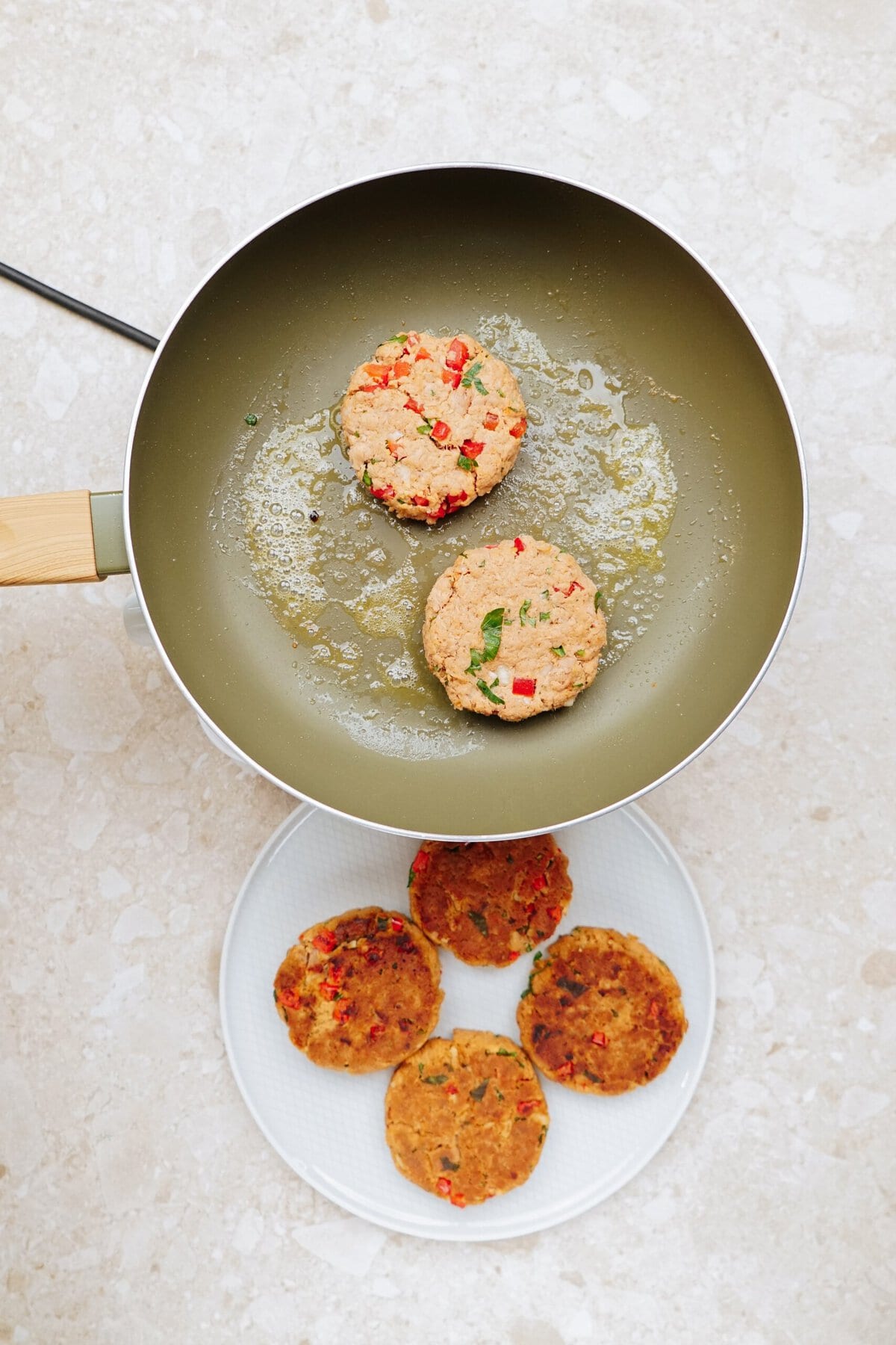 Two tuna patties sizzle in a non-stick frying pan, while three cooked patties rest on a white plate beside the pan.