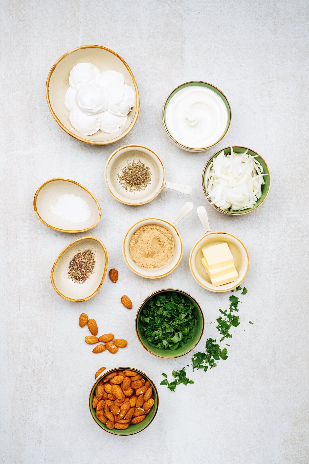 Overhead view of various ingredients in small bowls, including sour cream, yogurt, butter, ground spices, chopped herbs, onions, whole almonds, and salt. Nearby, a plate of goat cheese balls adds a touch of sophistication to the array.