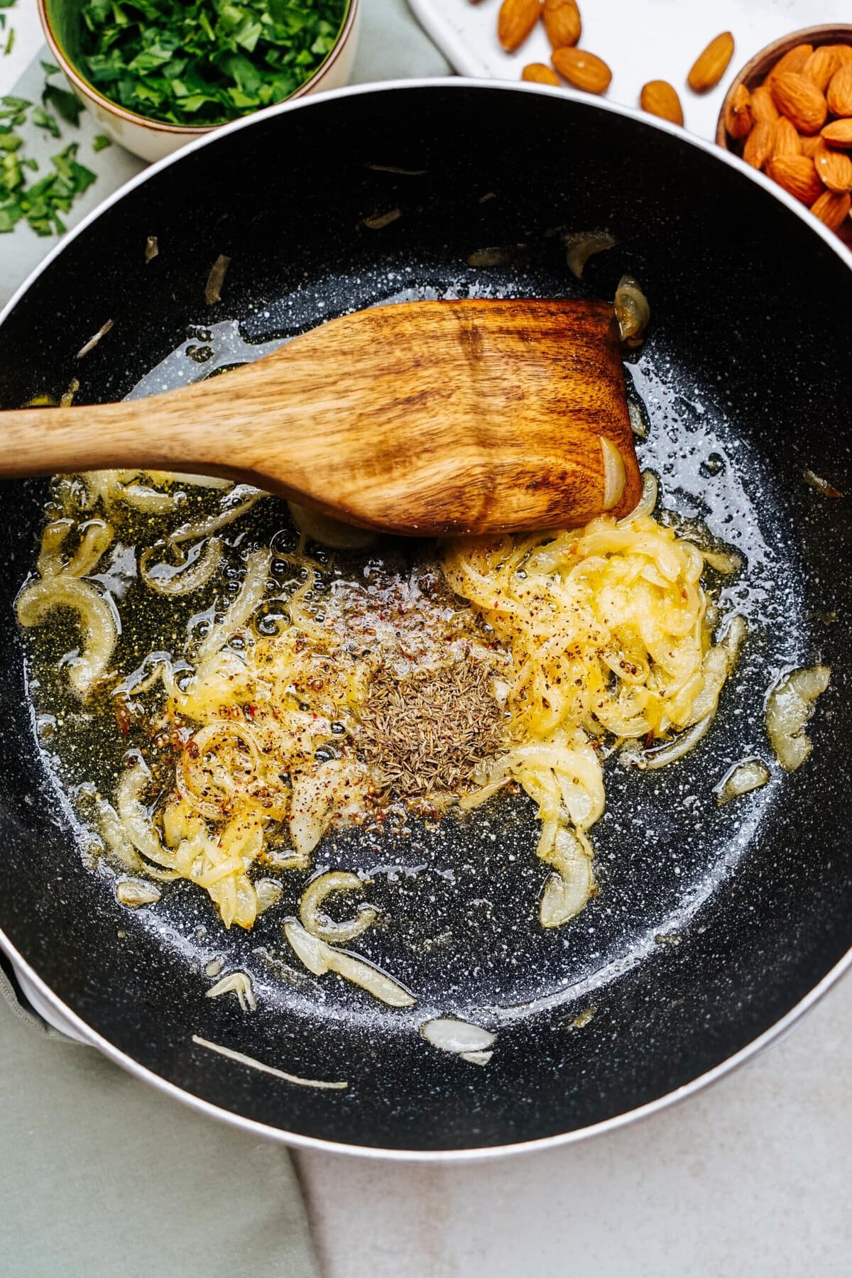 Cooking process in a frying pan with onions, spices, and a wooden spatula. Chopped herbs, almonds, and goat cheese balls are visible nearby.