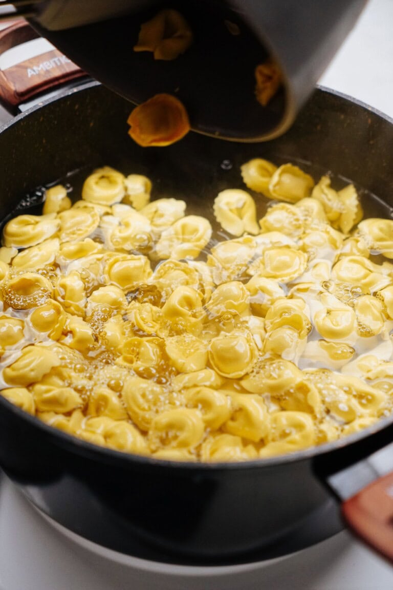 A close-up of yellow tortellini being poured from a pot into a black pan filled with boiling water.