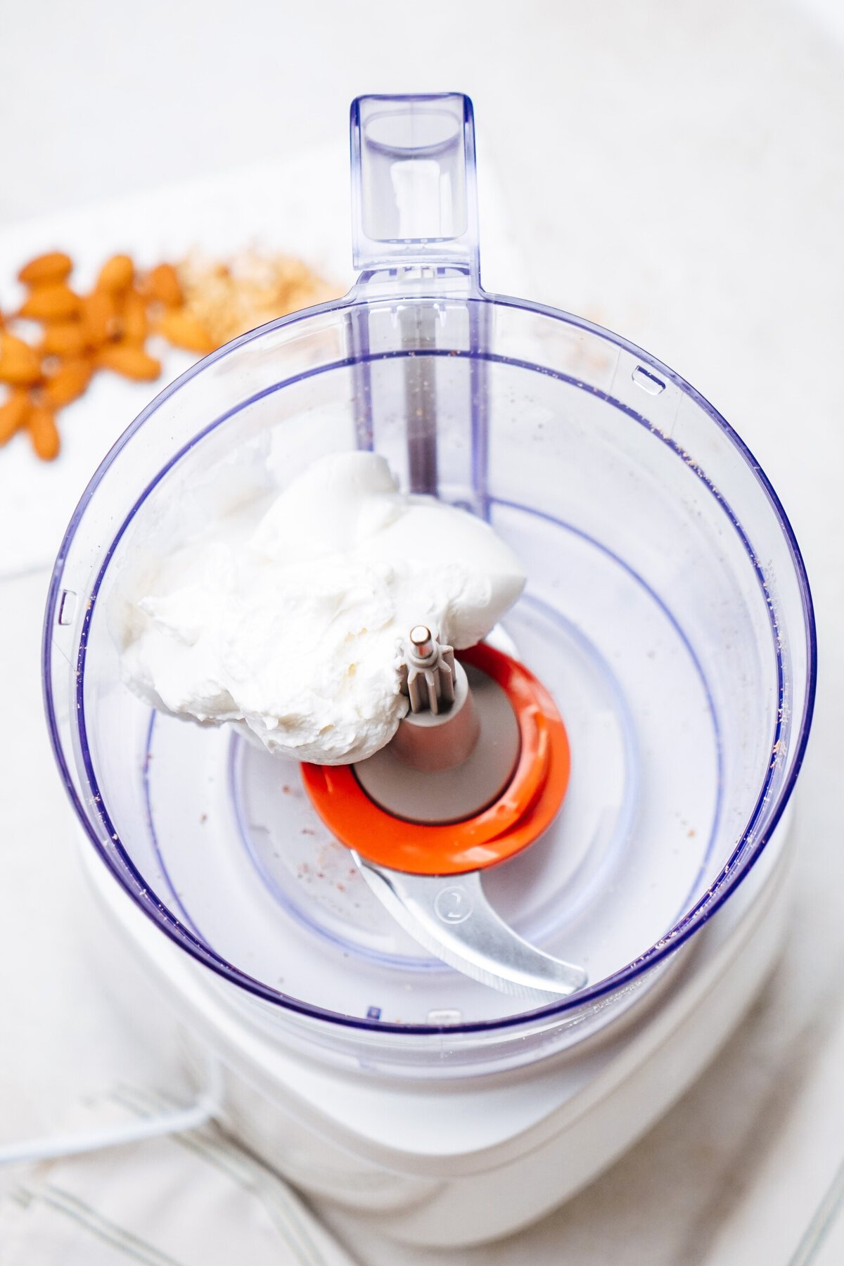 A food processor contains a mixture of white ingredients, presumably cream or whipped product, with a red and silver blade in the center. In the background, there are almonds and other small food items, perhaps for making decadent goat cheese truffles.