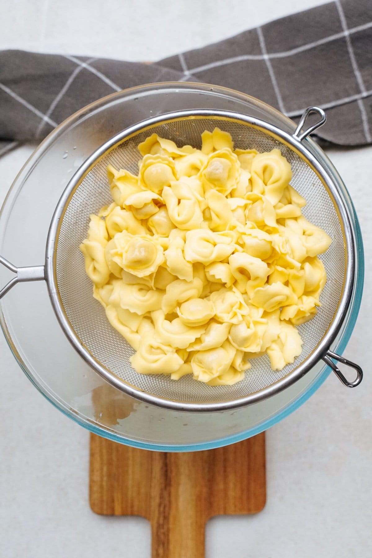 Cooked tortellini draining in a metal colander set over a glass bowl, with a gray checkered kitchen towel in the background.