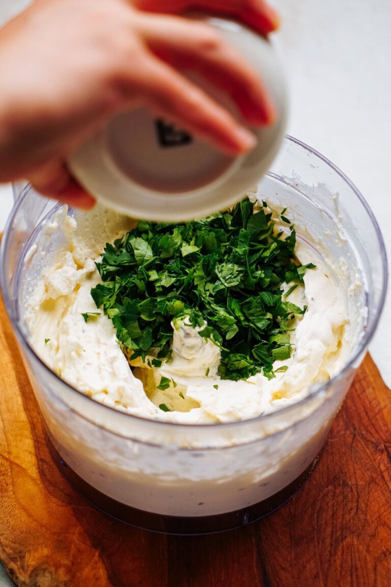 A person adds chopped fresh herbs to a food processor filled with a creamy mixture, crafting the perfect base for delectable goat cheese truffles.