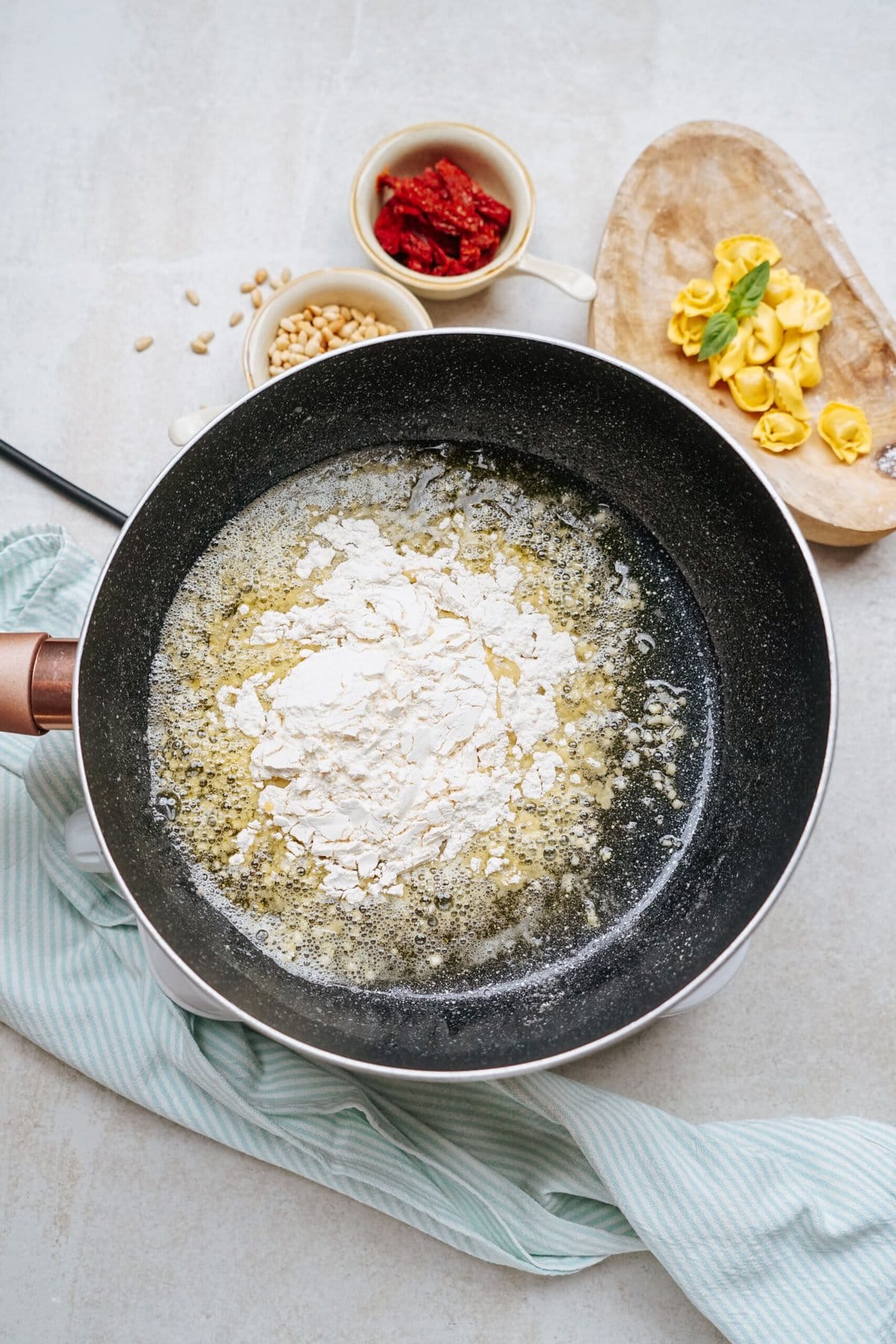 A frying pan with butter and flour. Small bowls of pine nuts and sun-dried tomatoes, and a wooden board with stuffed pasta are nearby.