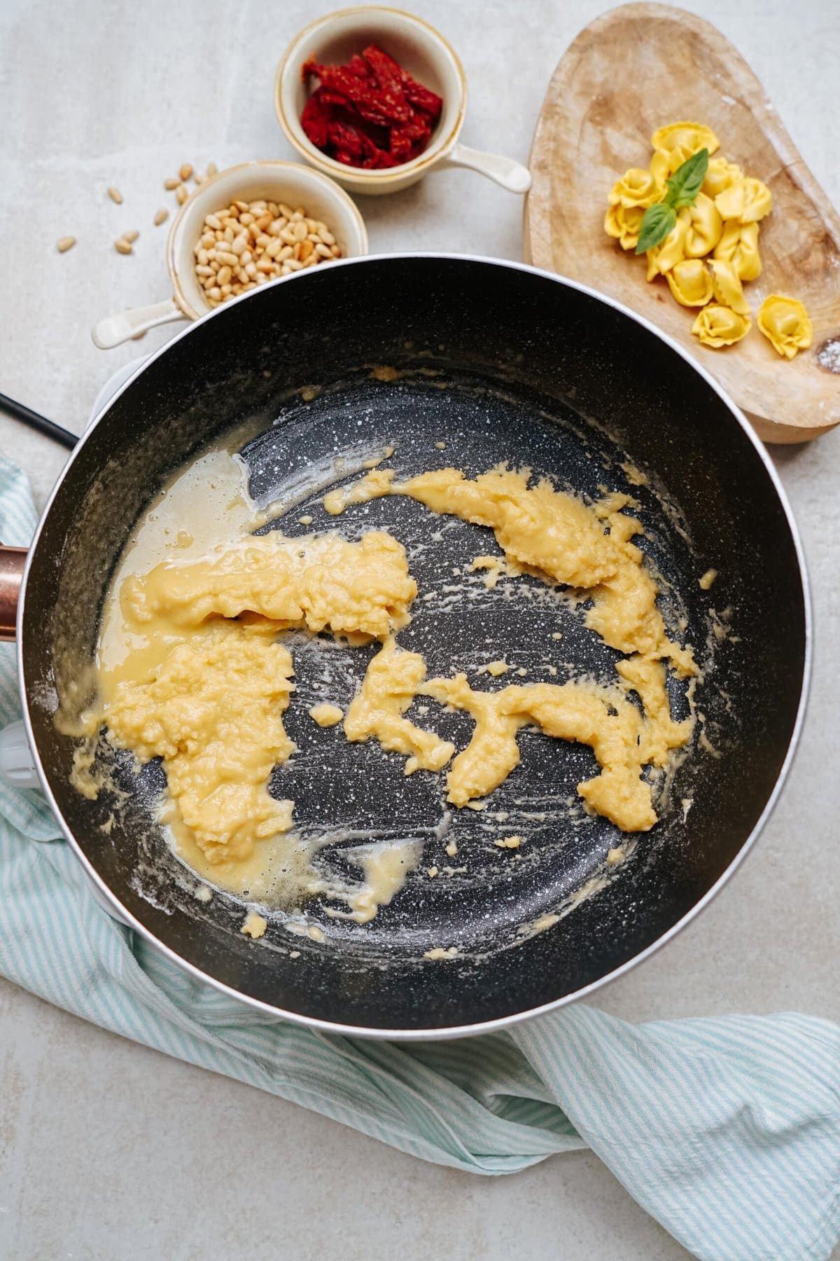 A pan with remnants of a yellow sauce sits on a kitchen counter. Nearby are dishes of pine nuts, sun-dried tomatoes, and uncooked tortellini on a wooden board.