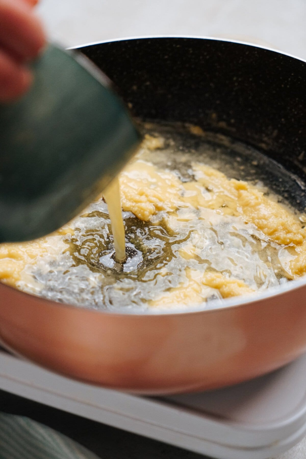 A person pours liquid from a green jug into a pan with cooking batter on a stovetop.