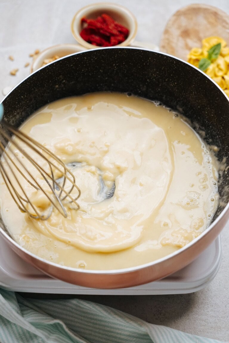 A saucepan containing a creamy white sauce being whisked, with bowls of ingredients and a cutting board in the background.