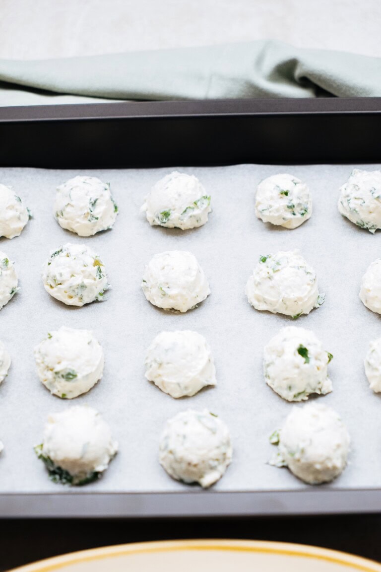 A baking sheet lined with parchment paper holds several scoops of a cheese and herb mixture, including delightful goat cheese truffles, prepared for baking.