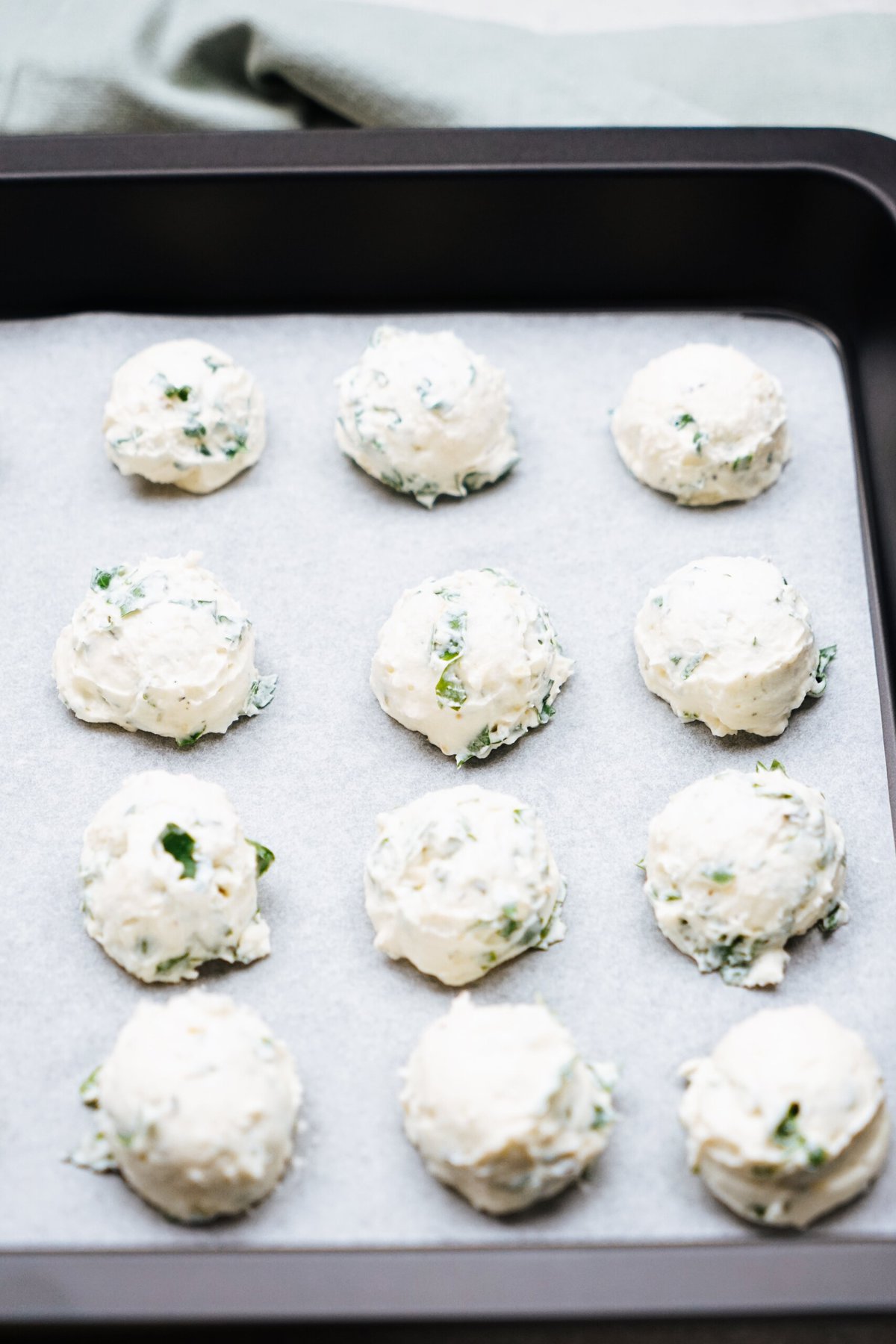 A baking tray lined with parchment paper holds twelve evenly spaced scoops of an uncooked ricotta herb mixture, reminiscent of goat cheese truffles.