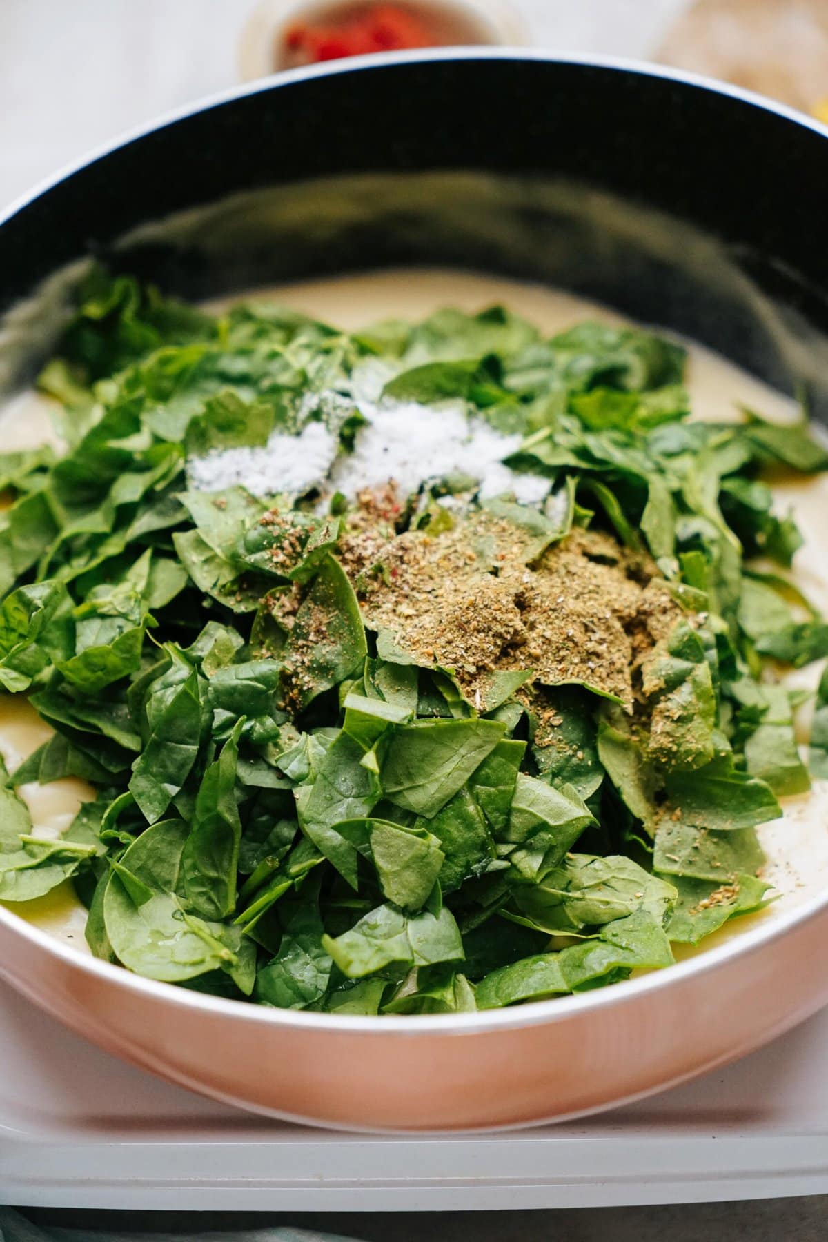 A pan containing chopped spinach, salt, and various spices being prepared, presumably before being cooked.