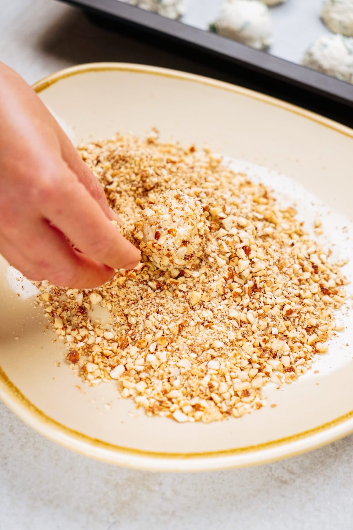 A hand is rolling a round goat cheese truffle in a plate of crushed nuts, with more similar items on a tray in the background.