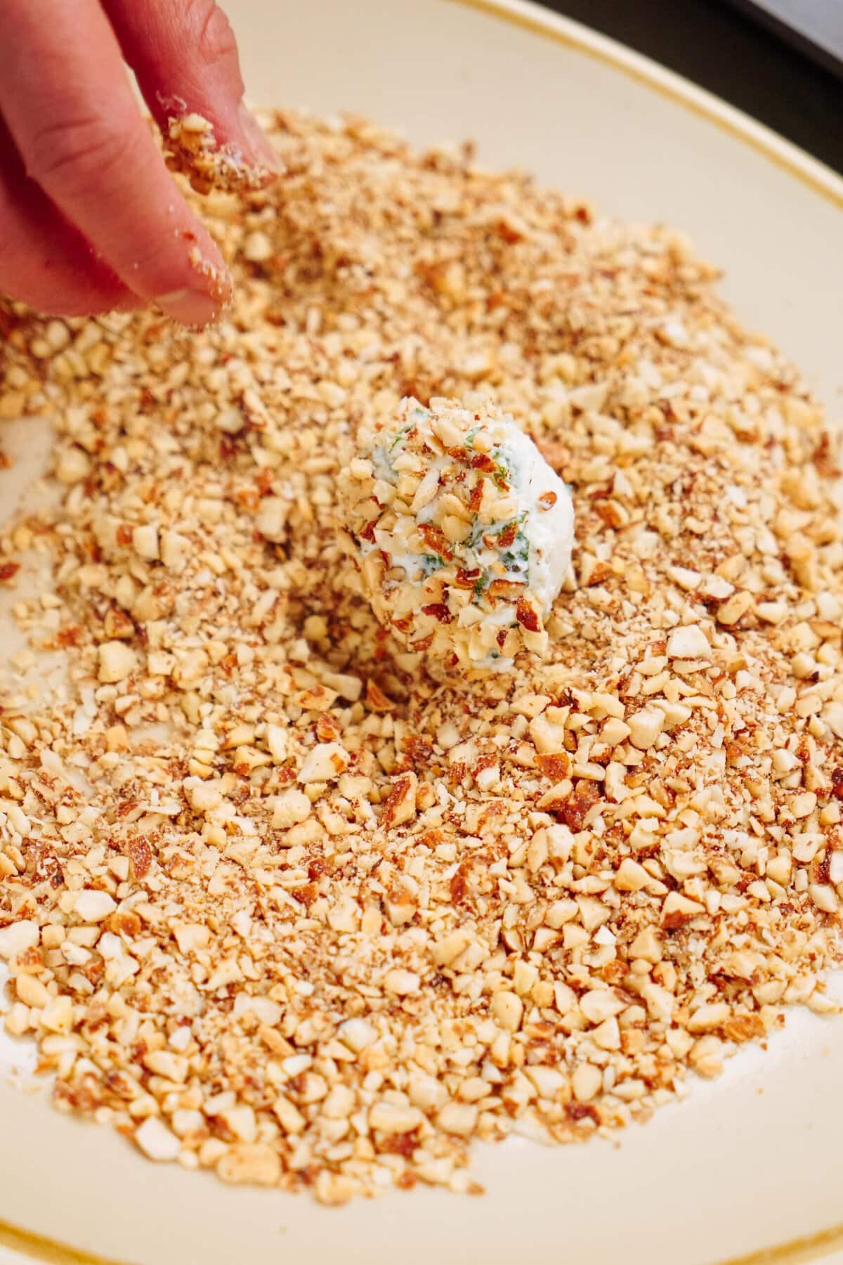 A hand is seen rolling a white, ball-shaped goat cheese truffle in a plate full of crushed nuts and herbs.