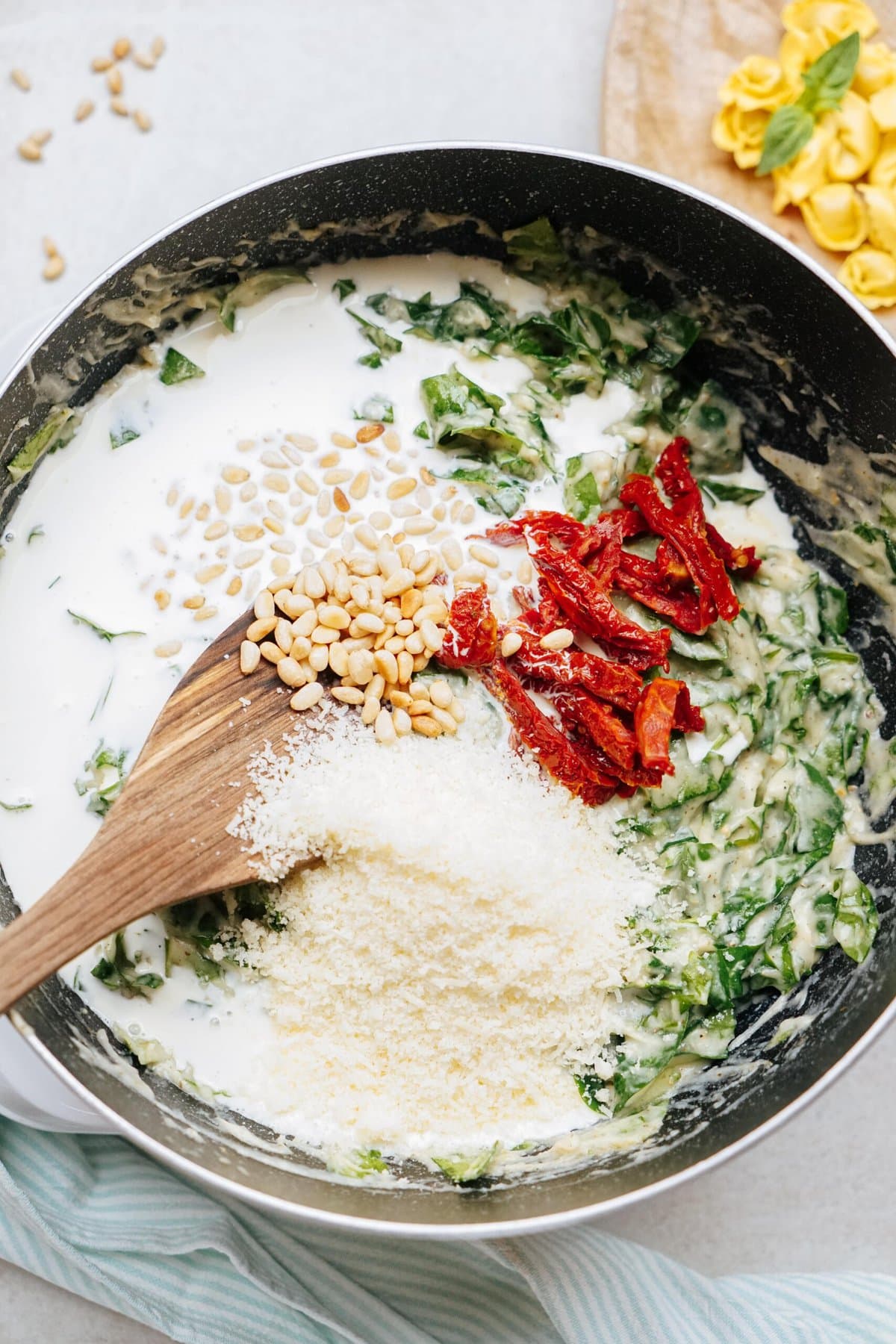 A skillet with a creamy sauce containing chopped greens, sundried tomatoes, pine nuts, and grated cheese. A wooden spoon rests in the skillet.