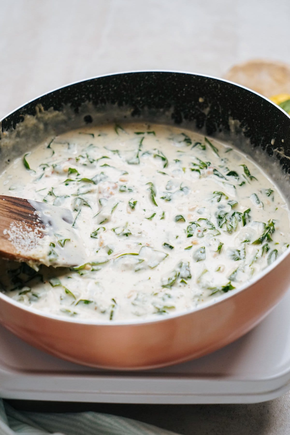 Close-up of a pan containing a creamy spinach dish being stirred with a wooden spoon. The creamy mixture is simmering on a stovetop.