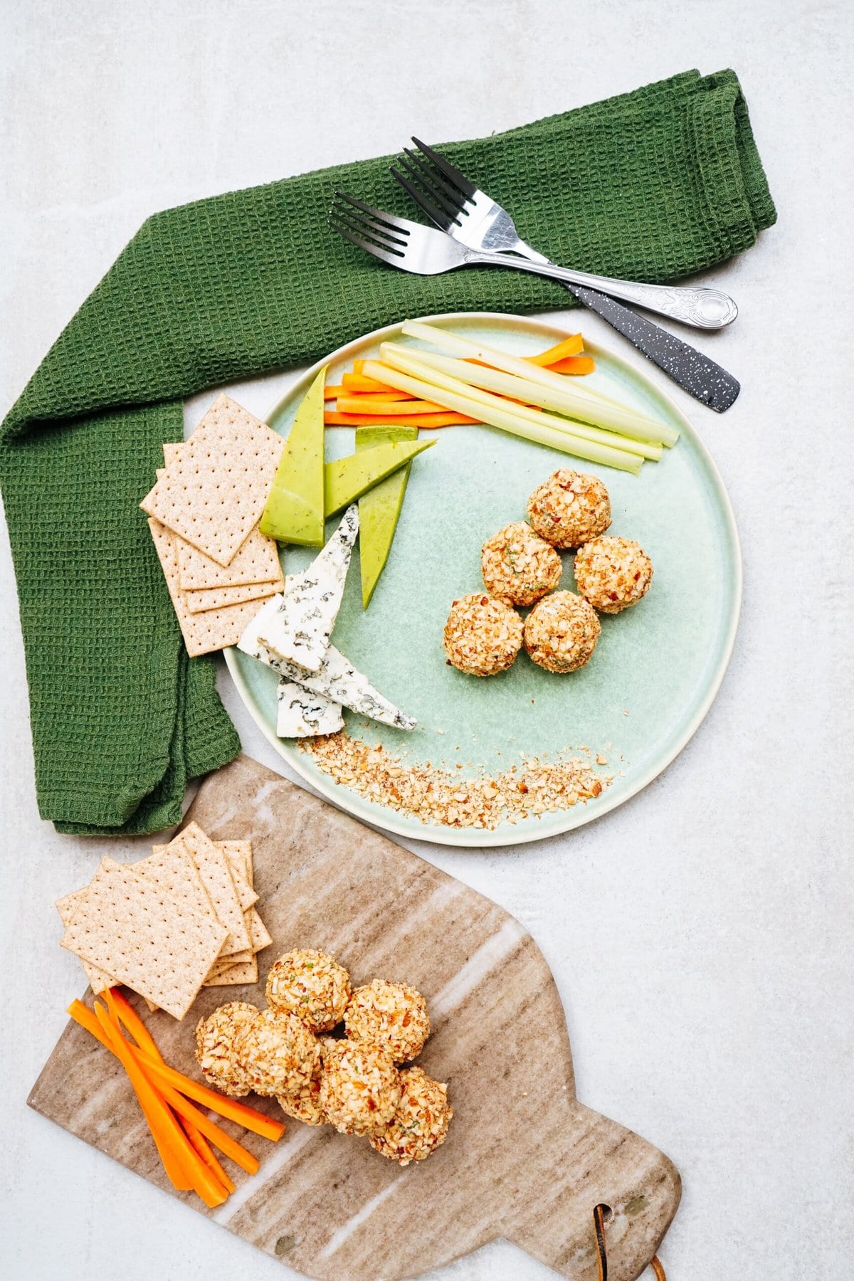 A plate of goat cheese truffles with herbs, slices of bread, celery, and carrot sticks next to a green cloth and utensils. Additional cheese balls and crackers are on a wooden board.