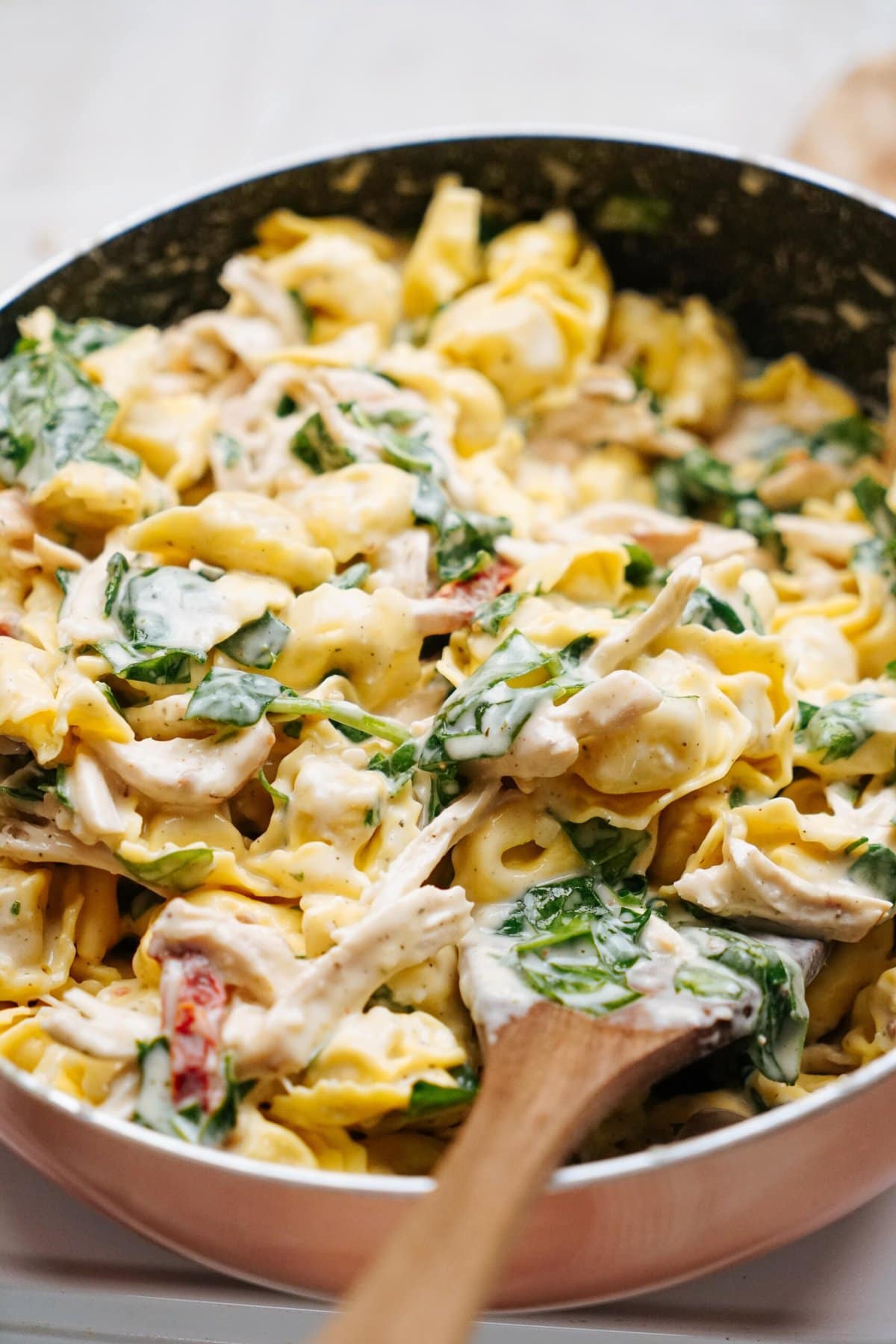 A skillet filled with creamy tortellini pasta mixed with shredded chicken, spinach, and sun-dried tomatoes, with a wooden spoon resting in the dish.