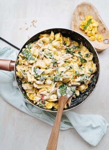 A skillet filled with creamy tortellini mixed with spinach and shredded chicken, with a wooden spoon inside and a small wooden board with additional tortellini and spinach in the background.