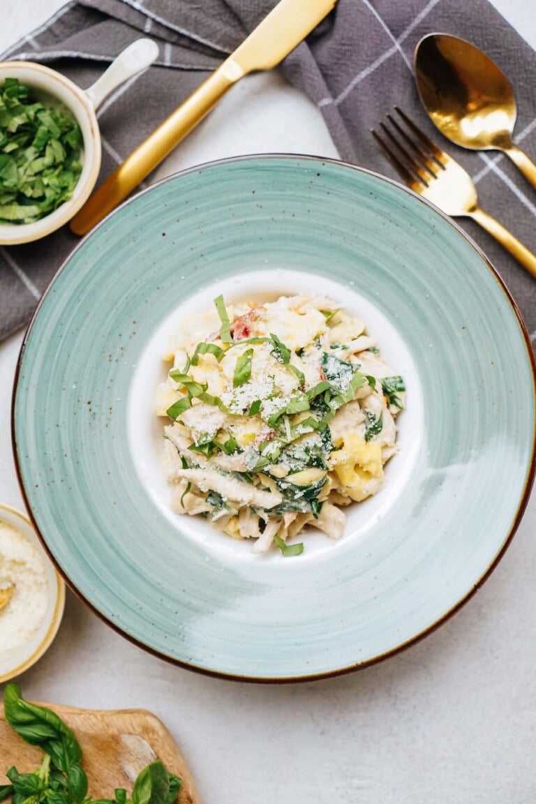 A plate of pasta topped with grated cheese and fresh herbs, placed on a table with a bowl of greens, wooden cutting board, and gold cutlery set on a gray napkin in the background.