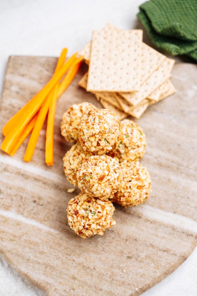Six goat cheese truffles coated in chopped nuts on a marble board, accompanied by thin carrot sticks, crackers, and a green cloth.