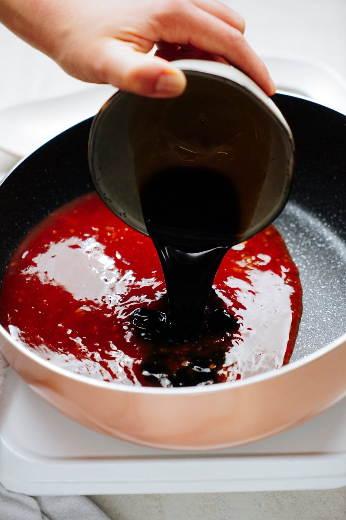 A hand pours a dark liquid into a frying pan containing a red sauce.
