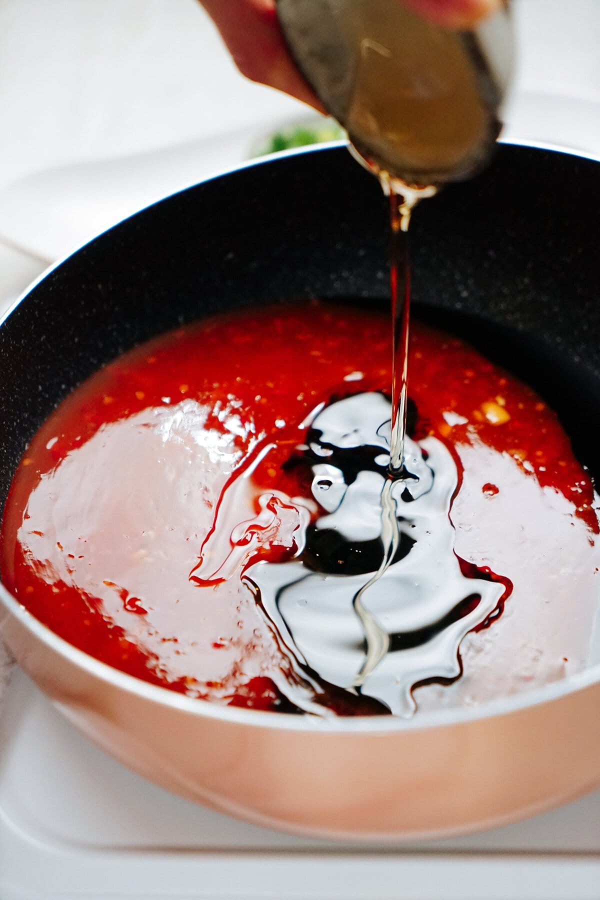 A hand is pouring a liquid ingredient into a pan filled with a red sauce.