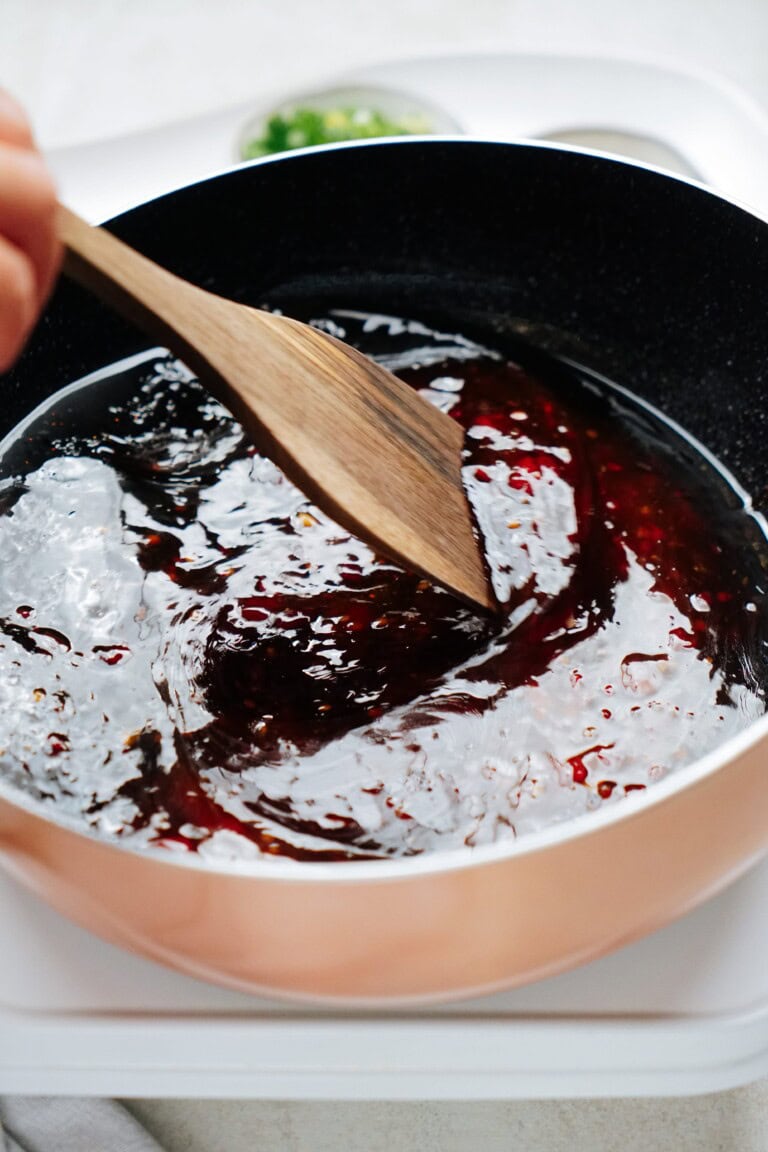 A wooden spoon stirring a dark red sauce in a black frying pan. The pan sits on a white surface, with some green garnish in a bowl partially visible in the background.