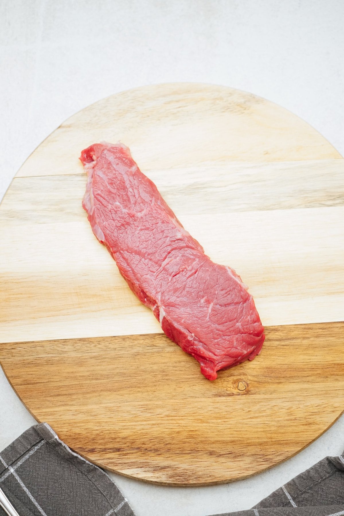 A raw piece of steak on a round wooden cutting board, with a gray and white kitchen towel partially visible at the bottom left.