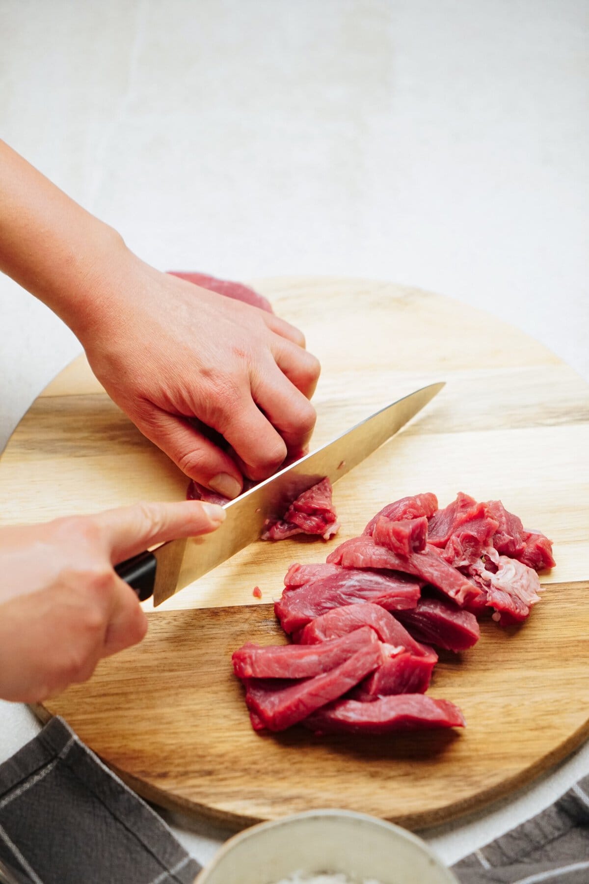 Close-up of a person slicing raw meat on a round wooden cutting board with a knife. A pile of sliced meat is also visible on the board.