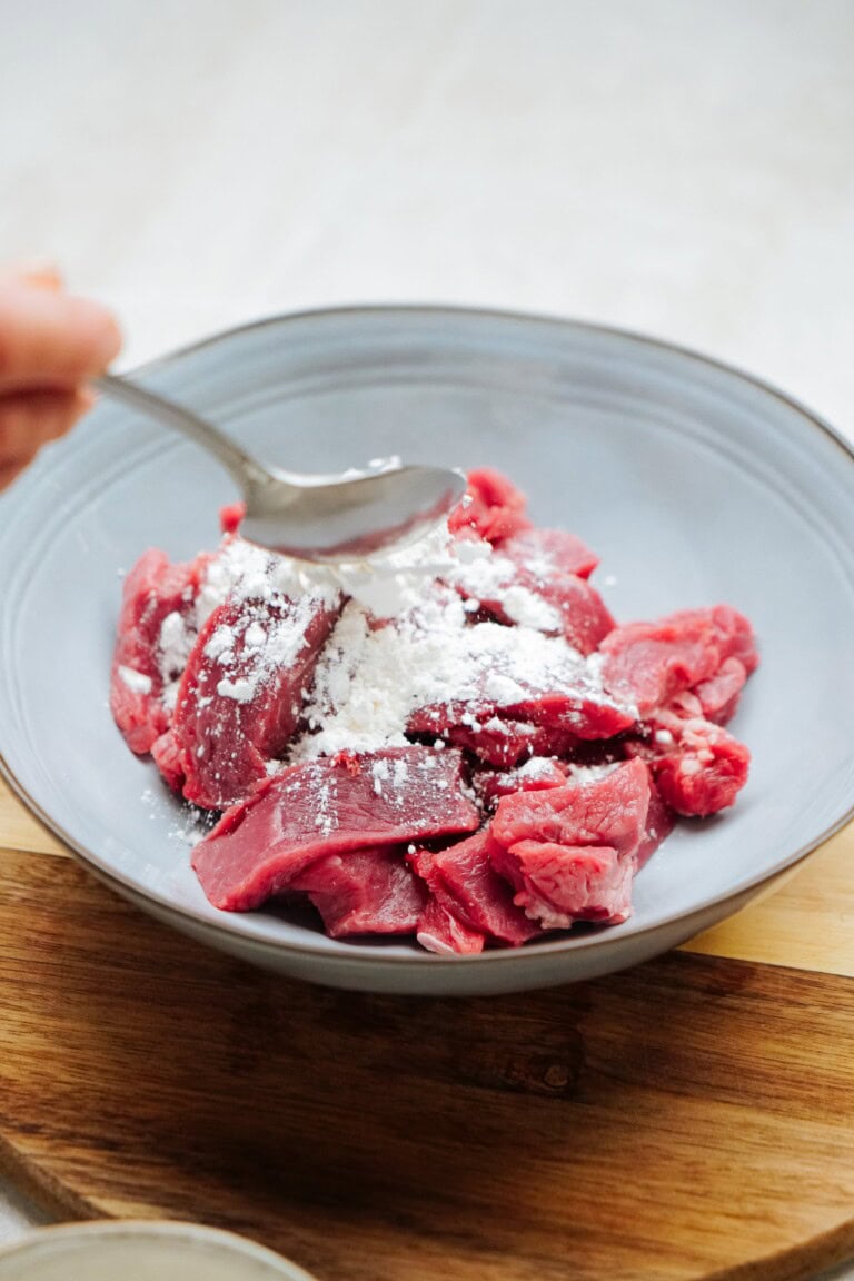 Close-up of raw beef strips in a bowl with white powder, likely flour, being sprinkled on top using a spoon.