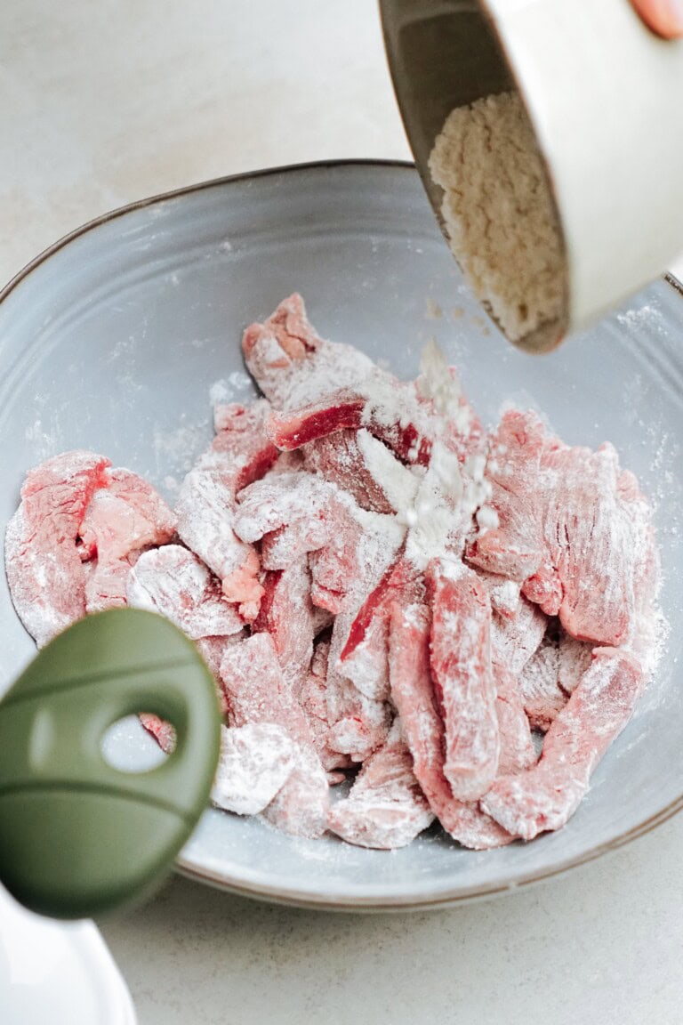 Raw beef strips coated in flour are in a bowl, with additional flour being poured from a container above. A green object is partially visible in the foreground.