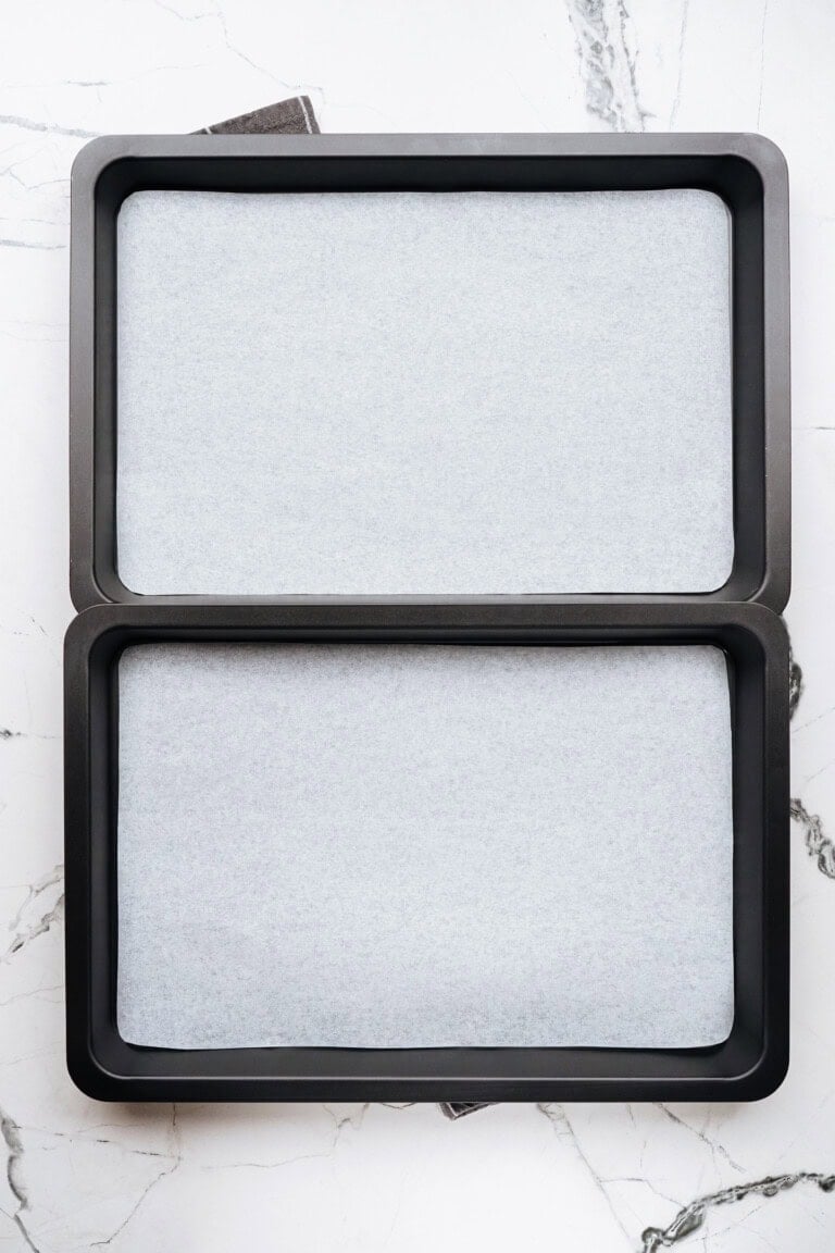 Two black rectangular baking trays lined with parchment paper, perfect for baking chocolate chip cookies, are placed side by side on a white marble surface.