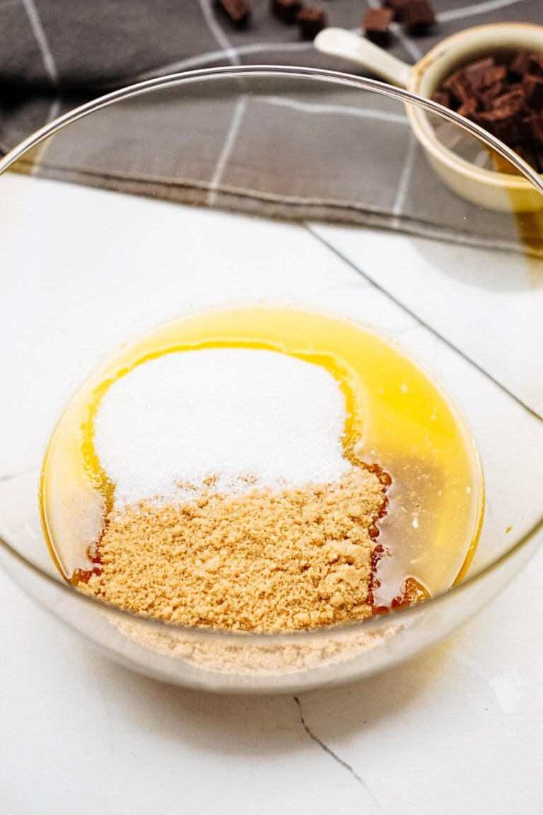 A glass mixing bowl containing unmixed ingredients like melted butter, granulated sugar, and brown sugar sits ready for action; in the background, a cutting board with chocolate pieces hints at delicious chocolate chip cookies in the making.