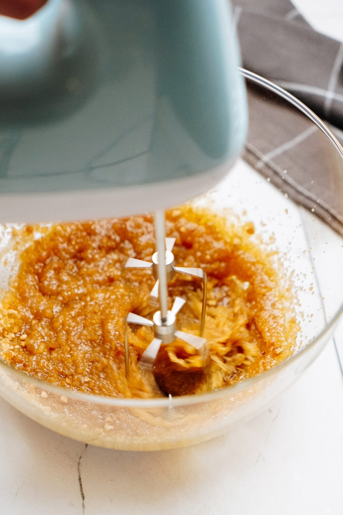 A blue hand mixer blends a brownish mixture for chocolate chip cookies in a clear glass bowl on a white surface.