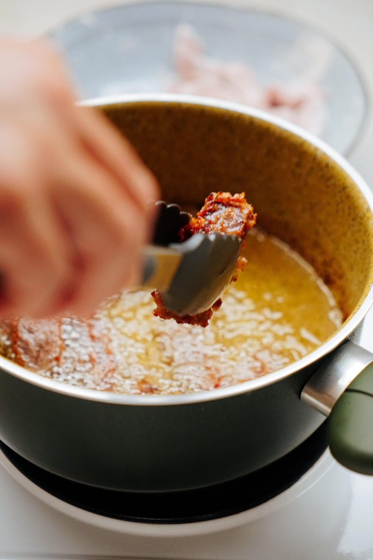 A hand holding tongs dips a piece of meat coated in breadcrumbs into hot oil in a frying pan on a stovetop.