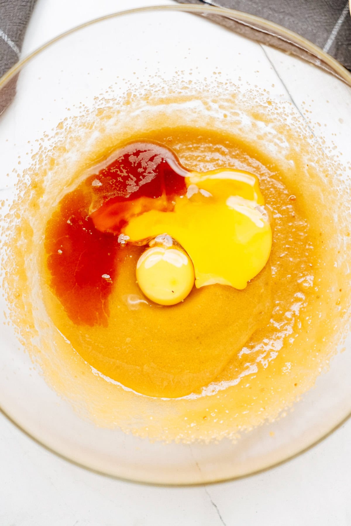 A glass bowl containing a mixture of egg yolk, vanilla extract, and a light brown batter, ready to become delicious chocolate chip cookies, rests on a white surface.