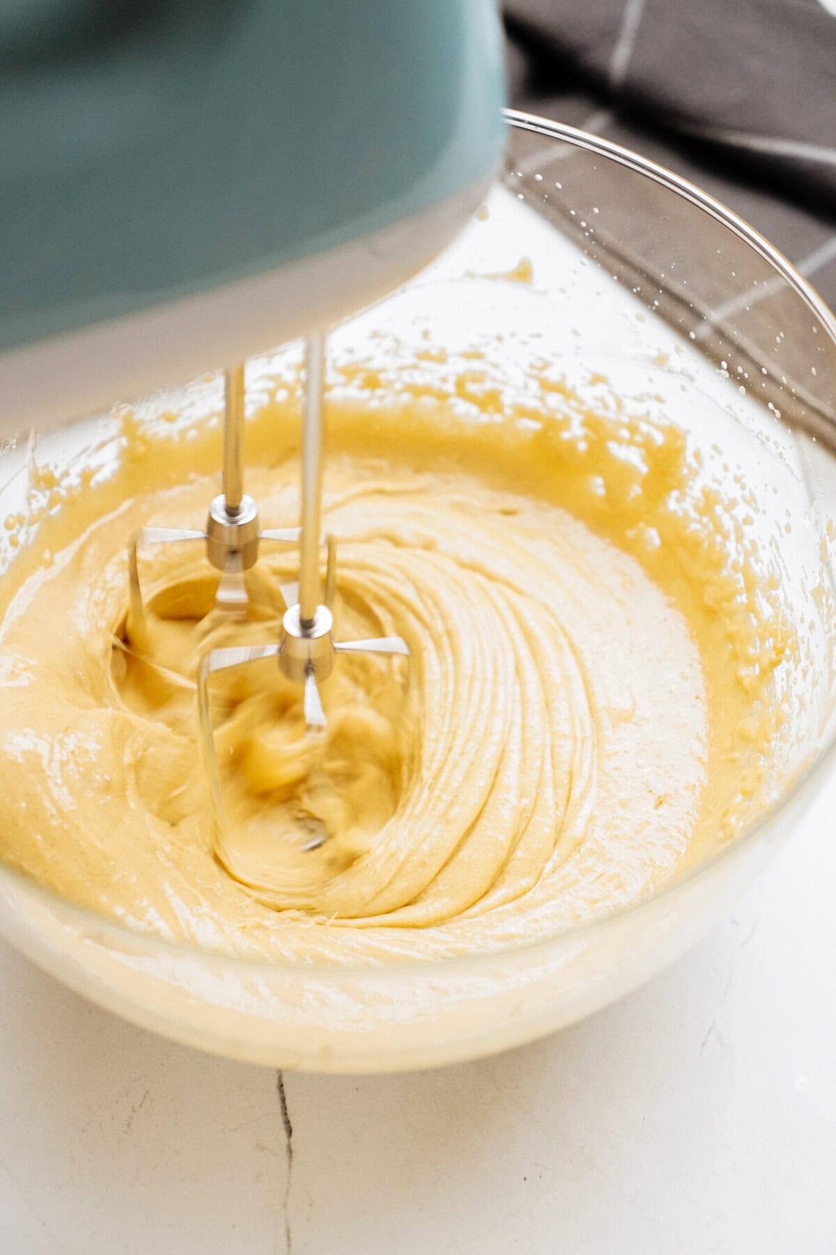 A close-up of chocolate chip cookie batter being mixed in a glass bowl with an electric hand mixer on a countertop. A brown cloth is partially visible in the background.