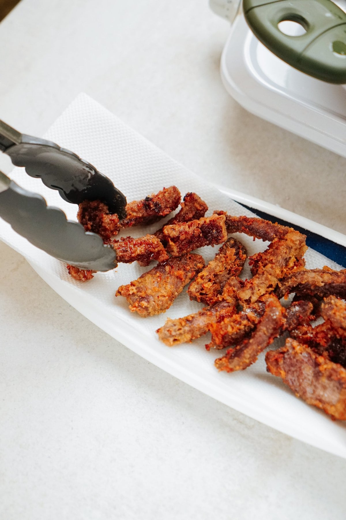 Tongs picking up pieces of fried beef from a paper towel-lined plate on a countertop, with a kitchen appliance in the background.