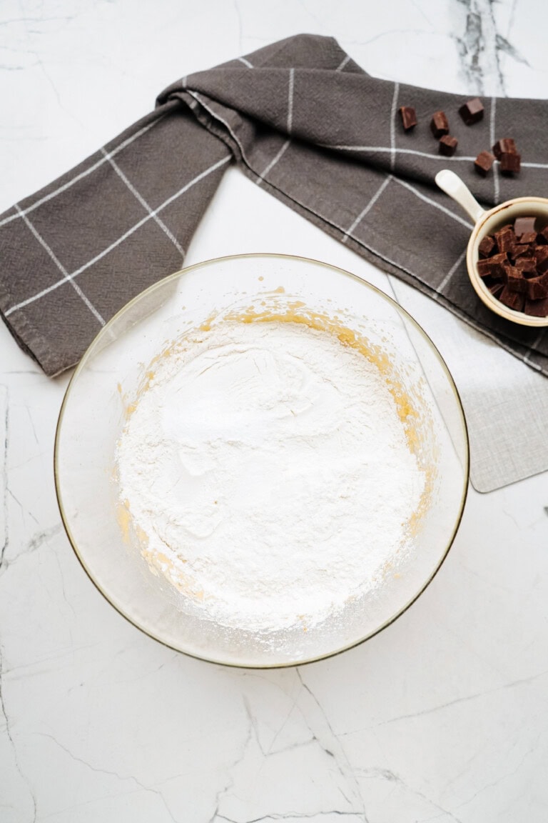 A glass bowl with flour and other ingredients on a marble countertop. Nearby, there is a folded gray cloth and a white measuring cup brimming with chocolate chunks, setting the stage for baking delicious chocolate chip cookies.