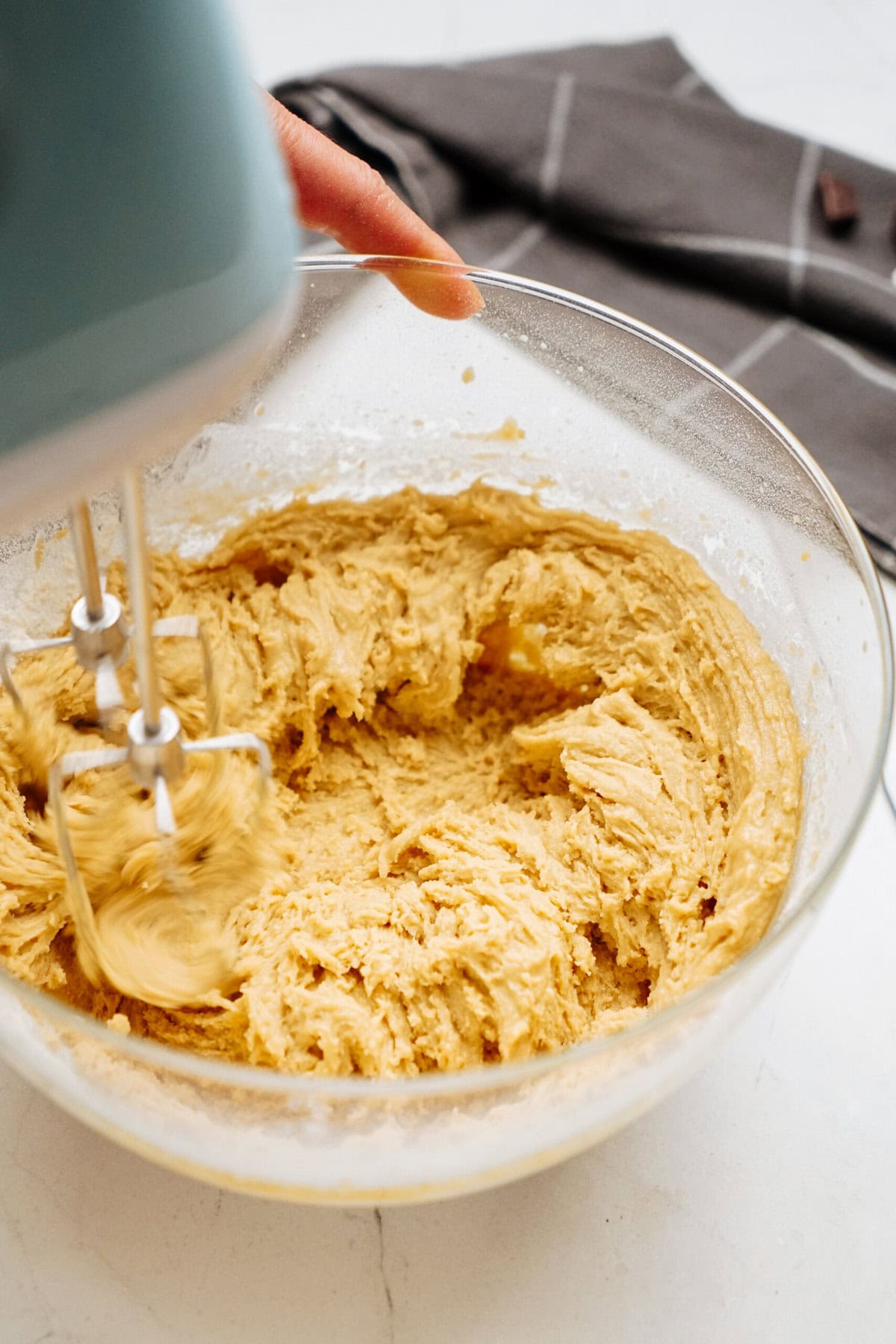 Hand holding an electric mixer creating a creamy dough mixture for chocolate chip cookies in a glass bowl, with a folded gray cloth in the background.