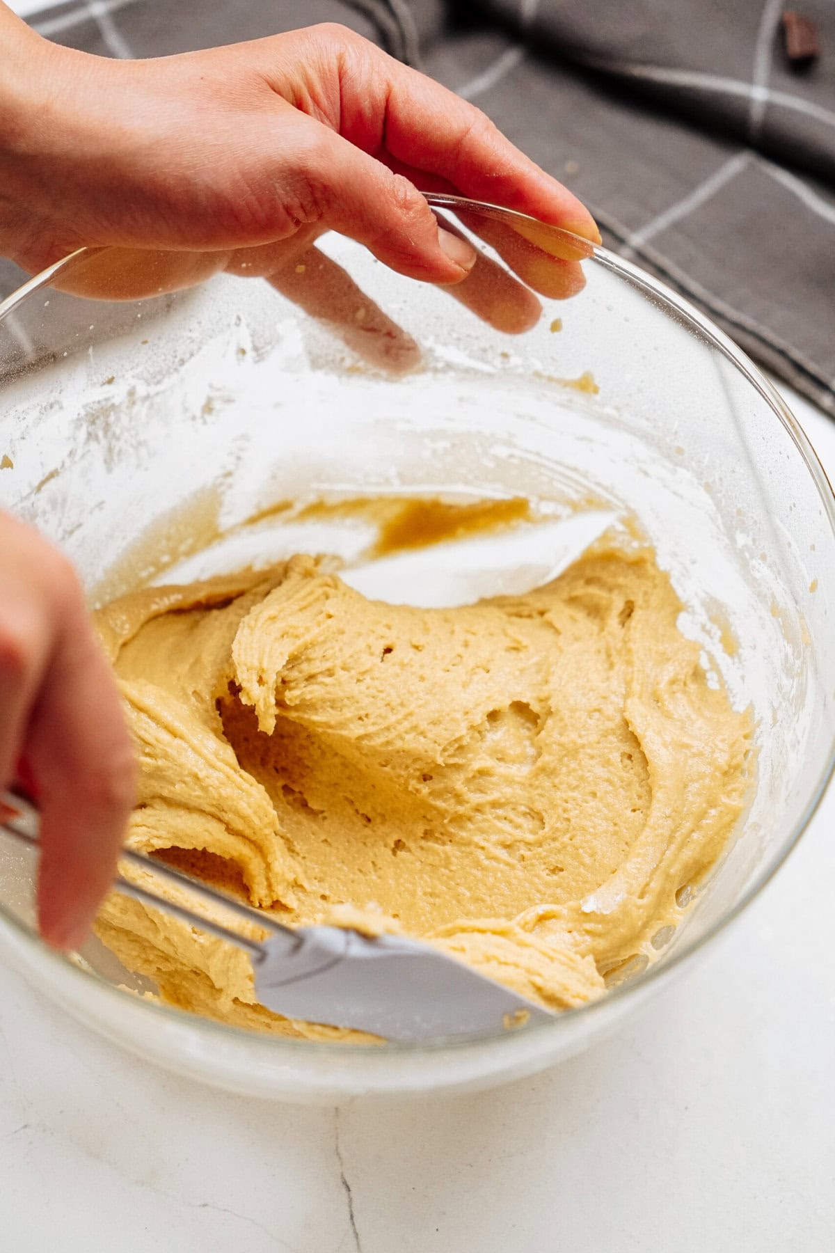 A person mixing a thick yellow batter for chocolate chip cookies in a glass bowl with a spatula.