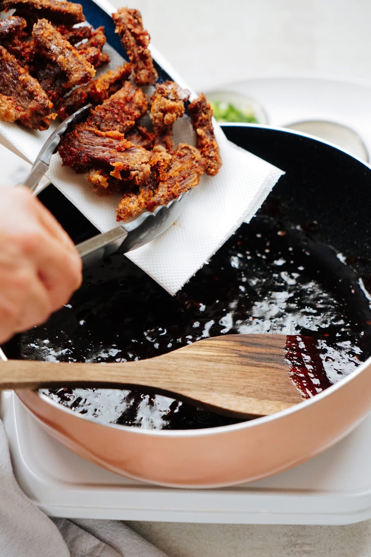 A person uses tongs to place fried beef onto a paper towel from a pan filled with oil. A wooden spatula rests in the pan.