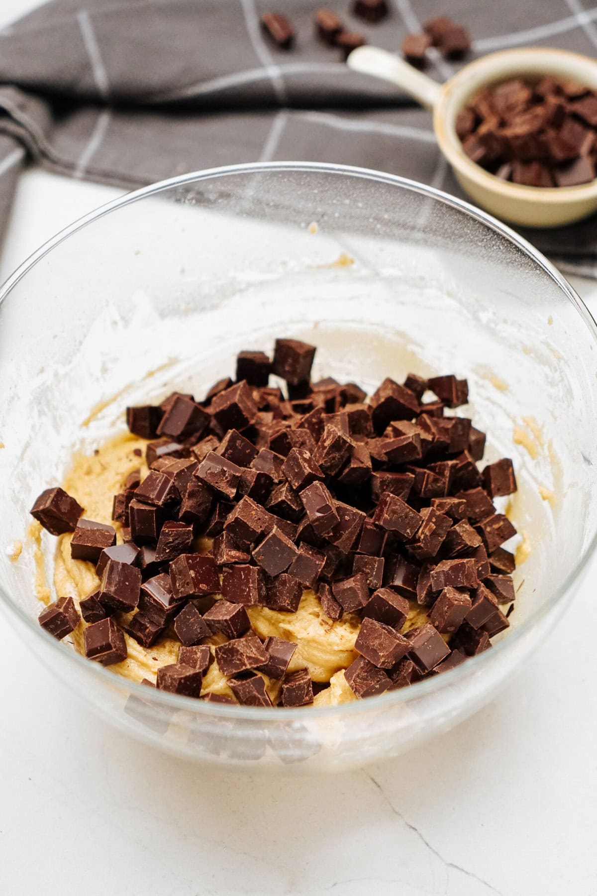 A clear glass mixing bowl containing cookie dough with chocolate chunks on top, ready to become delicious chocolate chip cookies. A small dish with more chocolate chunks and a gray cloth are in the background.