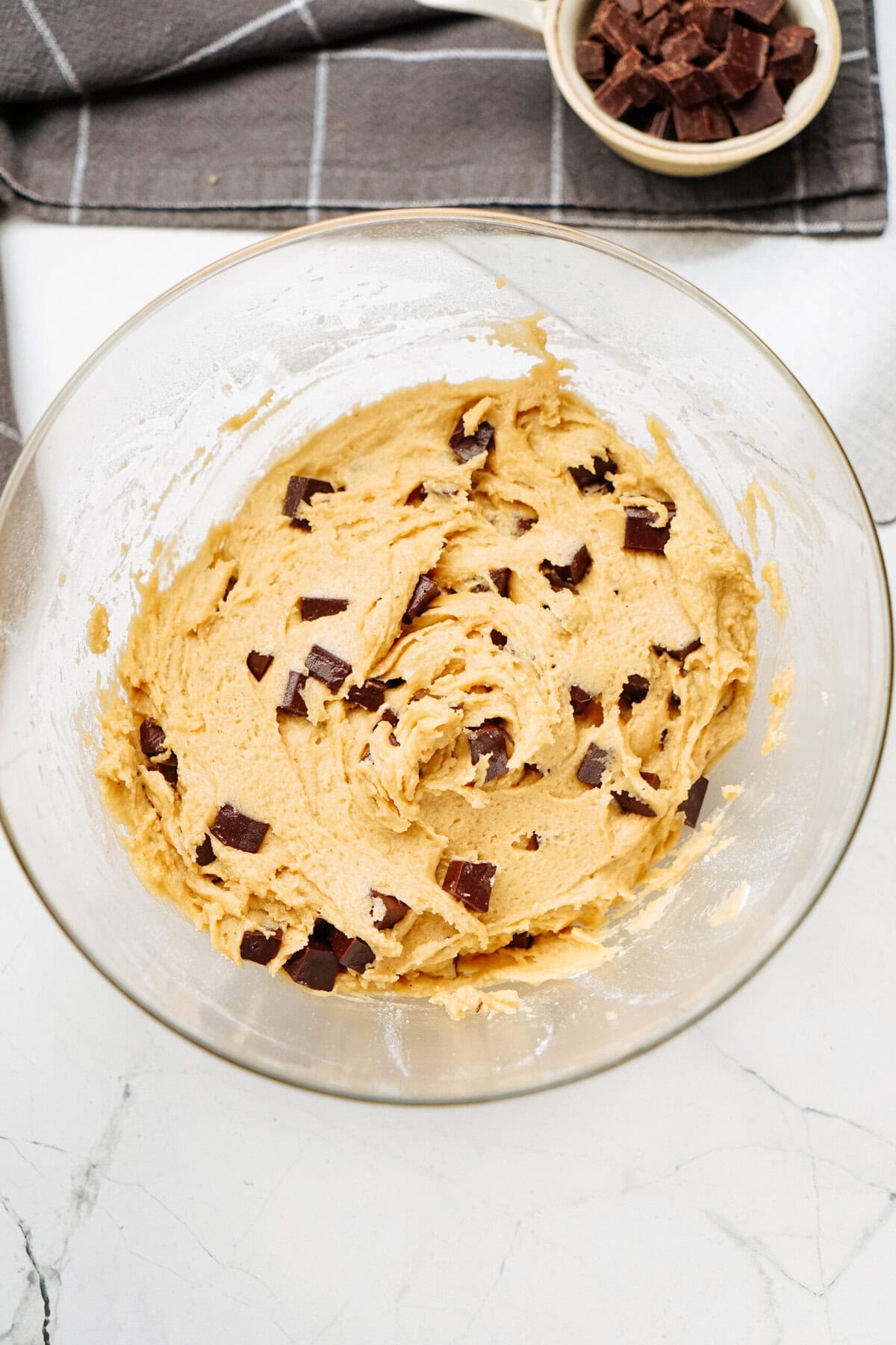 A glass bowl filled with cookie dough mixed with chunks of chocolate on a marble surface, promising the start of warm chocolate chip cookies. A small bowl of extra chocolate chunks waits in the background.