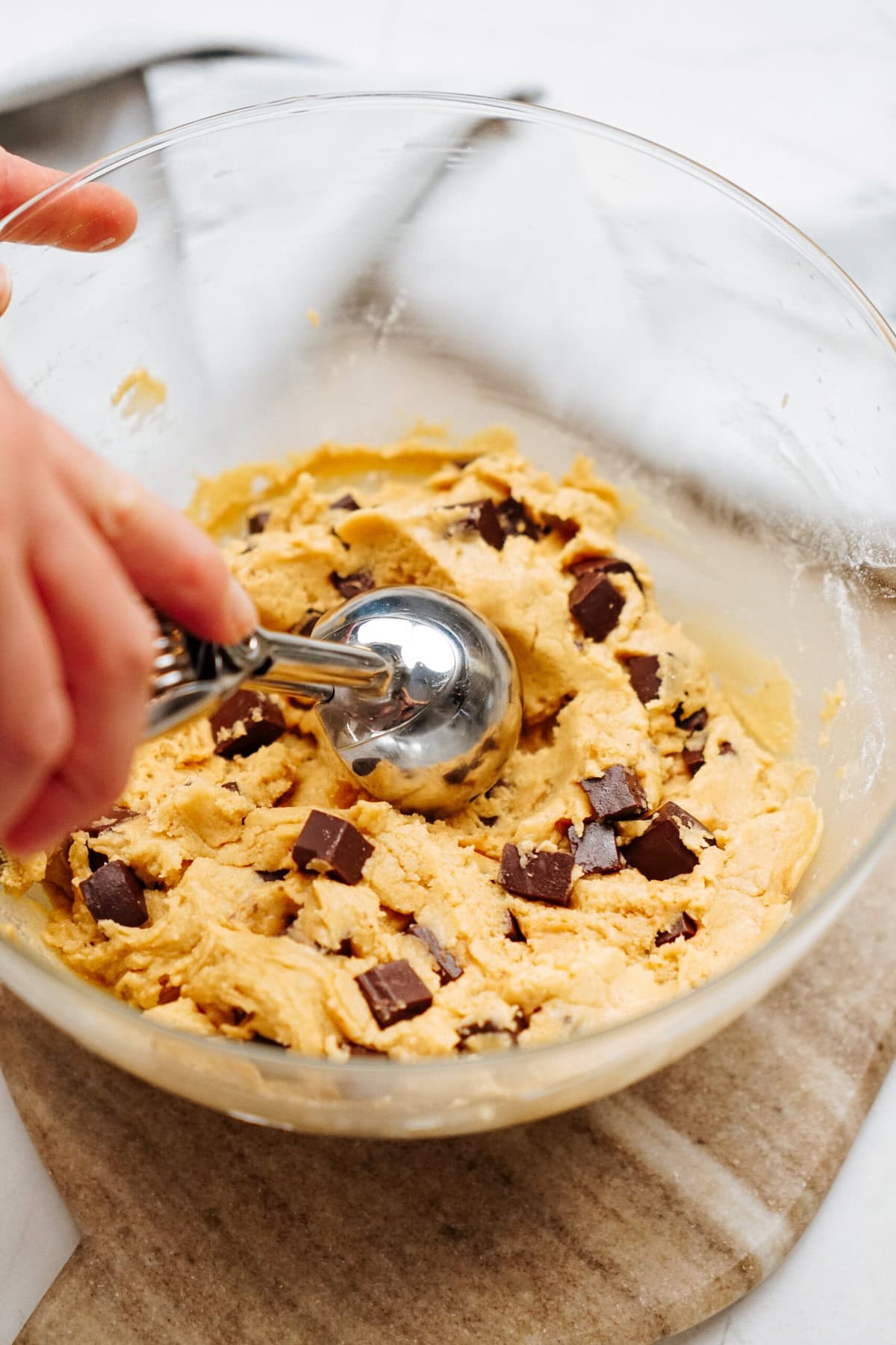 A hand uses a metal scoop to portion chocolate chip cookie dough with chocolate chunks from a glass bowl.